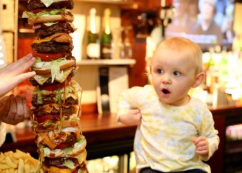 One year old Isla, who inspired Craig Harker's attempt at the UK's tallest burger looks bemused as she sits on the bar next top the 28 inch high meal. See SWNS story SWBURGER; A restaurant owner has created the daddy of all meals - Britain's tallest burger which is bigger than his DAUGHTER. The whopping two-and-a-half-foot '999 burger' is higher than Craig Harker's toddler and trumps the previous UK record by several inches. It gets its name from the 30,000 calorie content - enough to feed one person everyday for TWO WEEKS - which could leave any diner dialling for an ambulance. Regulars at The George in Stockton, Teeside, have been giving owner Mr Harker a big patty-on-the back for the 28 inch culinary masterpiece.