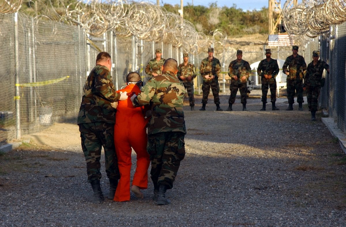 Two US Army (USA) Military Police (MP) escort a detainee, dressed in his new orange jumpsuit to a cell at Camp X-Ray, Guantanamo Bay Navy Base, Cuba. Camp X-Ray is the holding facility for detainees held at the US Navy (USN) Base during Operation ENDURING FREEDOM.
