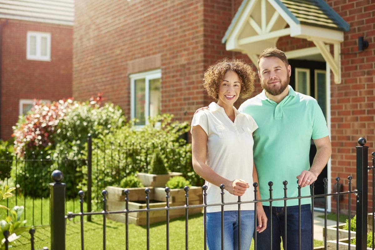 a mid adult couple stand at the front of their house and smile to camera . They are a mixed race couple , and their house is part of a new build housing development.