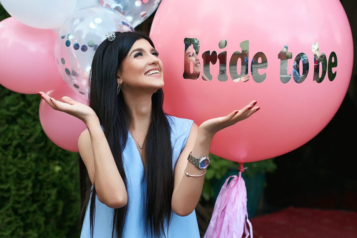 Happy young beautiful brunette bride to be with dark hair and silver crown smiling and gesturing spread out her hands in joy on bachelorette pink party balloon background.